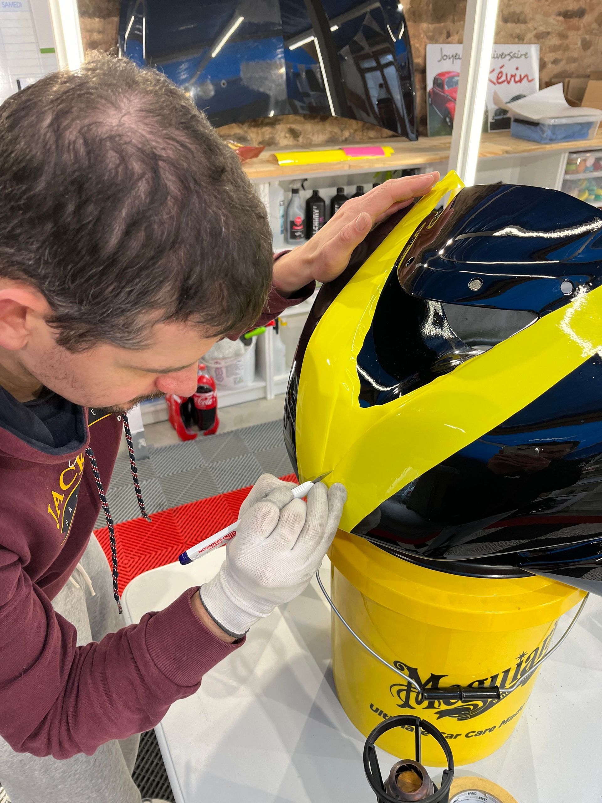 Une personne portant des gants peint une bande jaune sur un casque bleu et jaune posé sur un seau jaune.