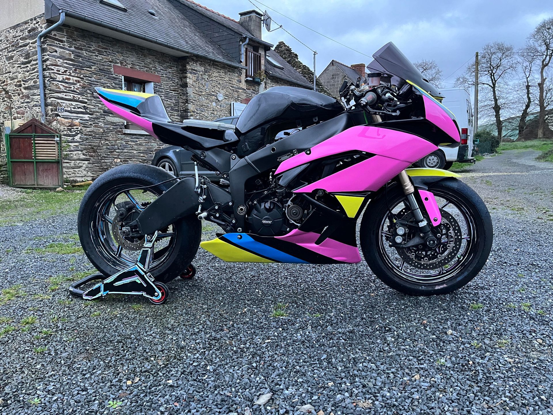 Moto de sport rose, noire et bleue garée sur une allée de gravier, avec un bâtiment en pierre en arrière-plan.