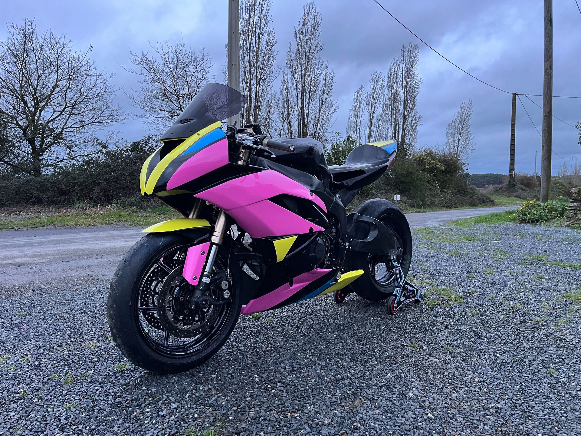 Moto de sport rose et jaune garée sur un chemin de gravier, journée nuageuse.
