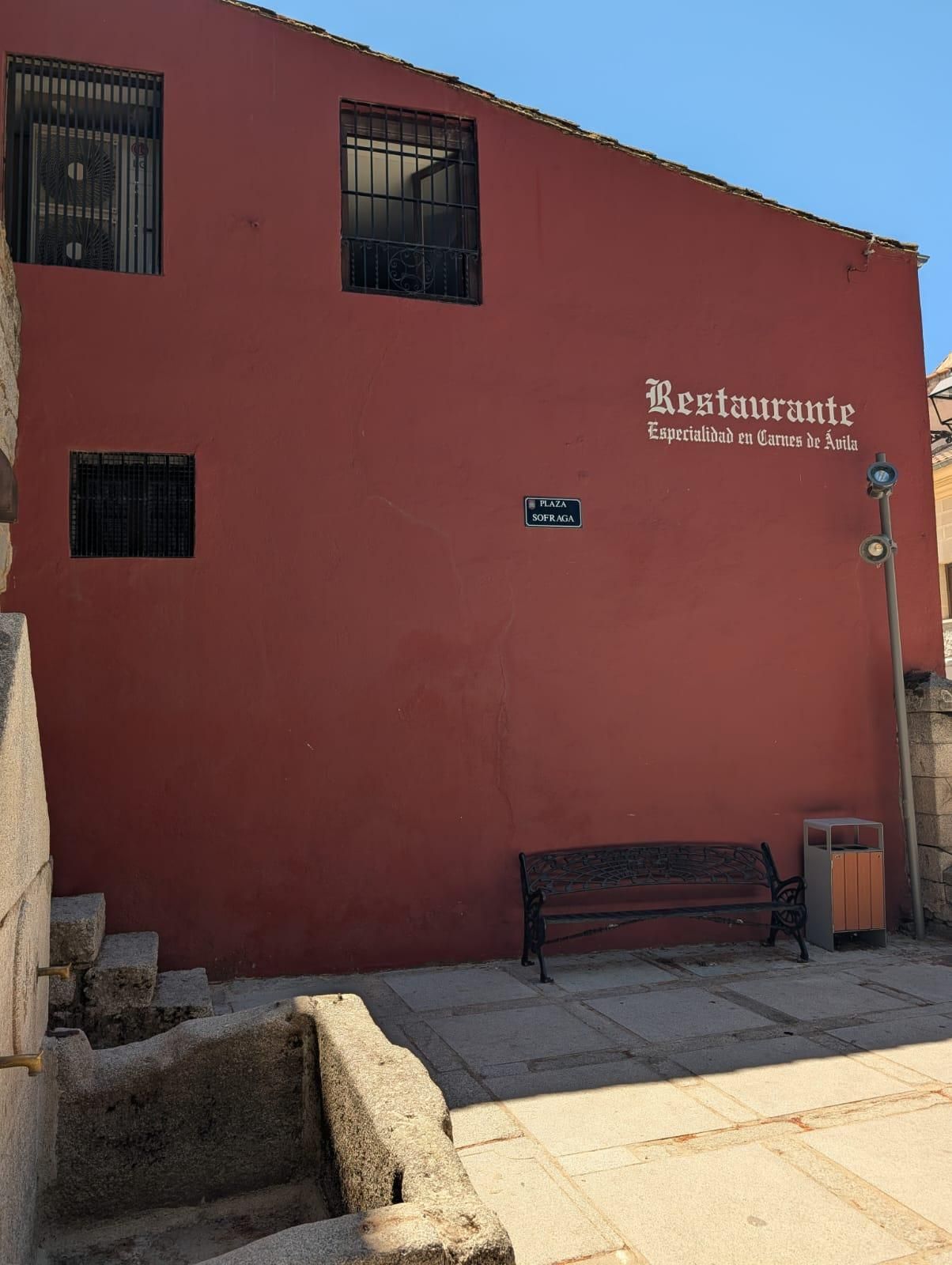 Fachada de restaurante de estuco rojo con pequeñas ventanas, banco y acera soleada.