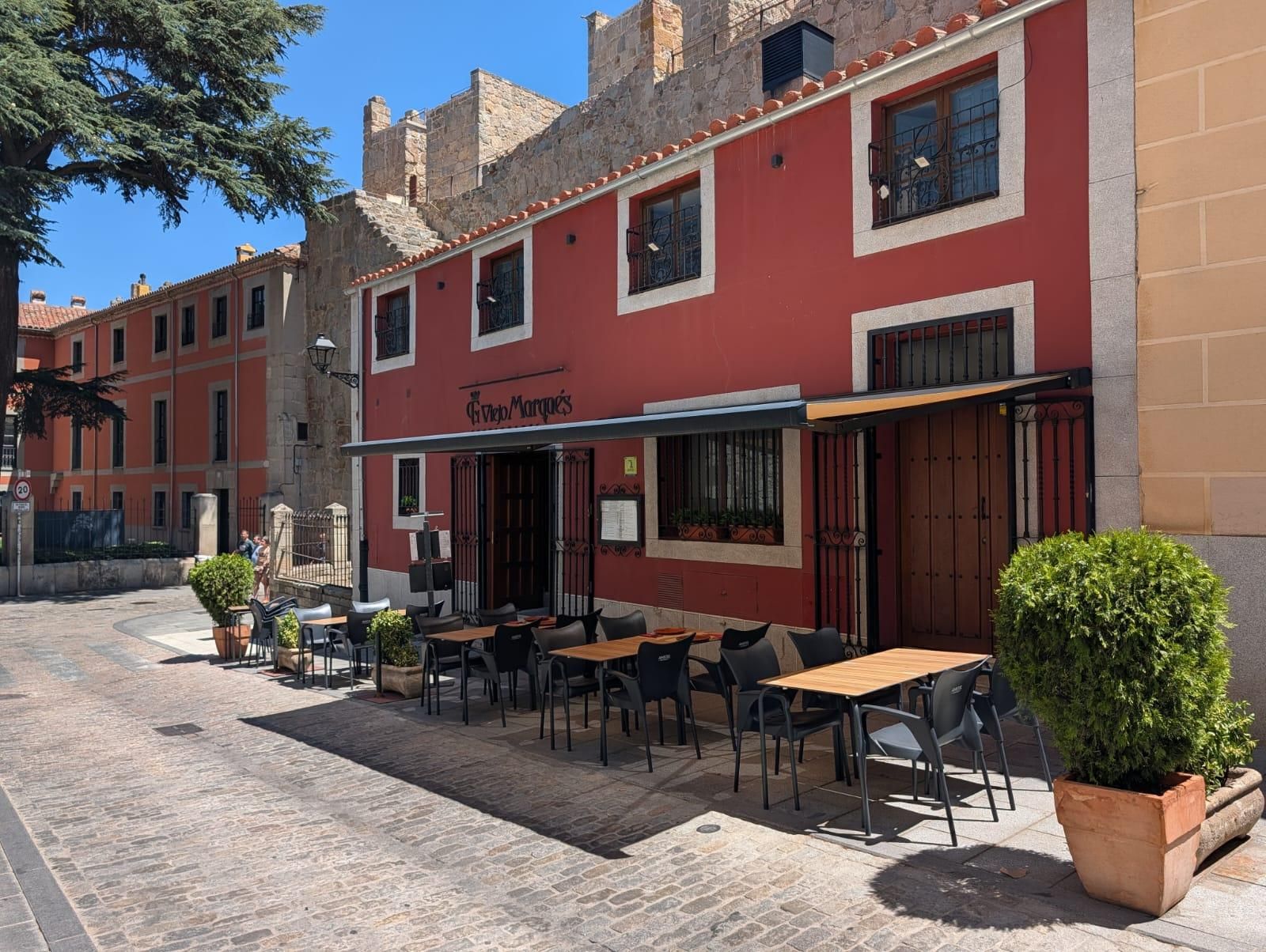 Cafetería al aire libre con fachada roja, mesas, sillas y arbustos en macetas en una calle empedrada.