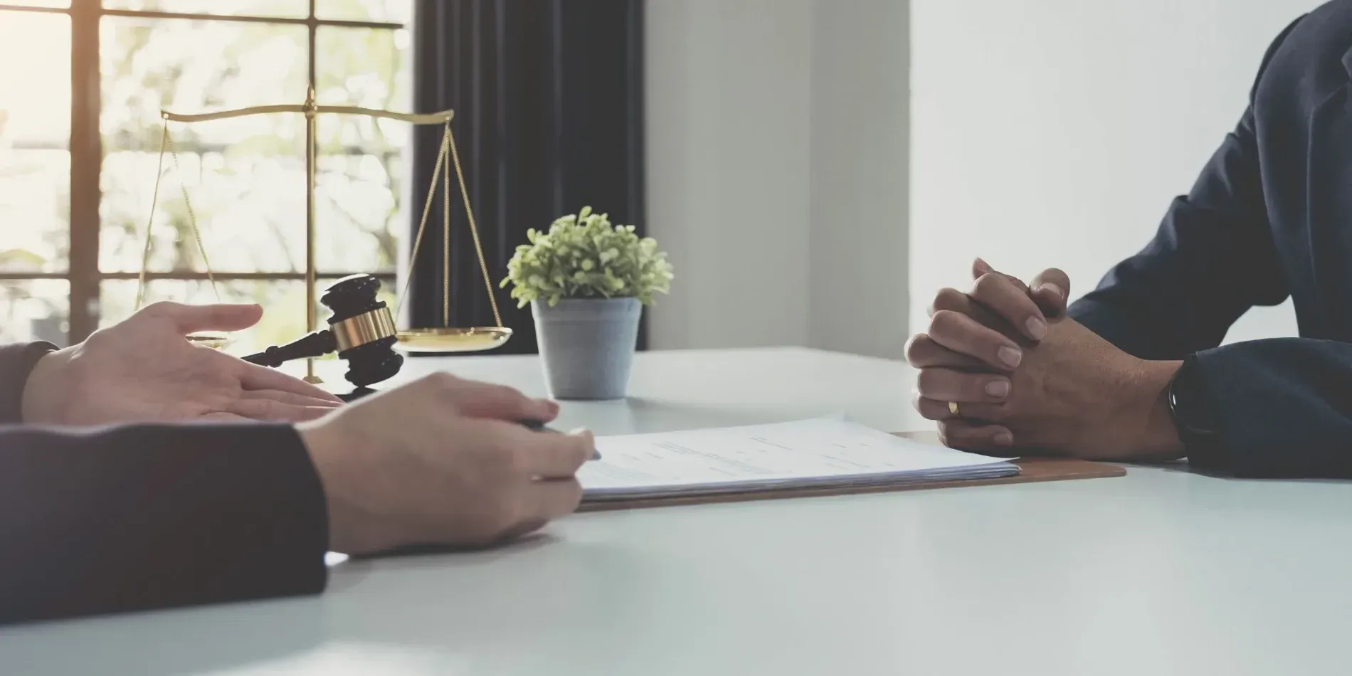 Dos personas de traje sentadas a una mesa; una gesticula mientras habla. Se ven un mazo, una balanza y una planta.