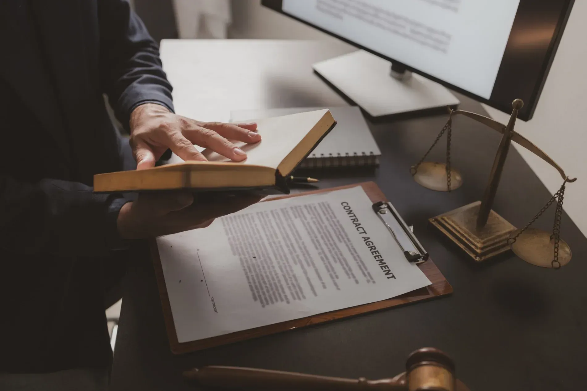Una persona examinando un libro cerca de un contrato, una balanza de la justicia, un monitor de computadora