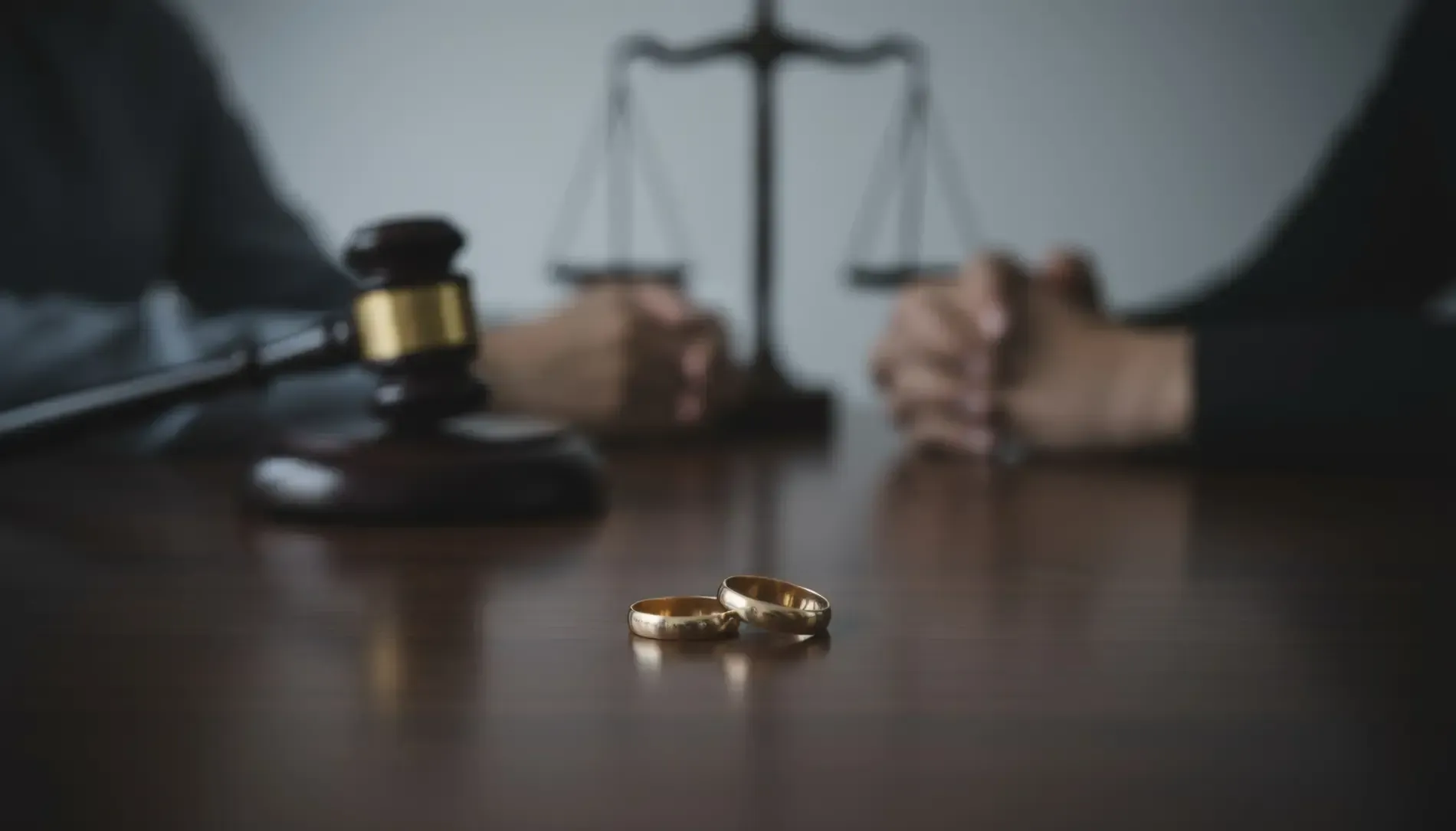 Anillos de boda sobre una mesa delante de un mazo y una balanza de la justicia; dos personas en el fondo.