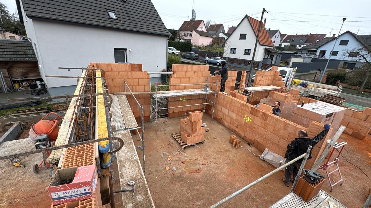 Fondations d'une maison en briques partiellement construite, avec des échafaudages et des maisons voisines sous un ciel couvert.