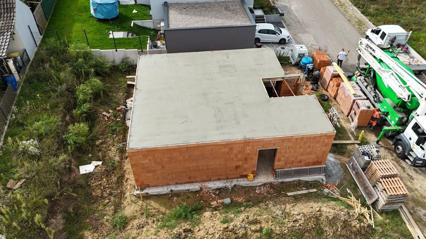 Vue aérienne d'un bâtiment en briques au toit gris clair, entouré d'engins de chantier et d'un terrain vague.
