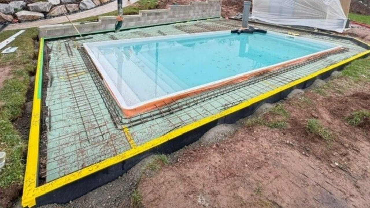 Piscine rectangulaire aux bordures jaune vif et orange, entourée d'un grillage, sur un terrain en pente.