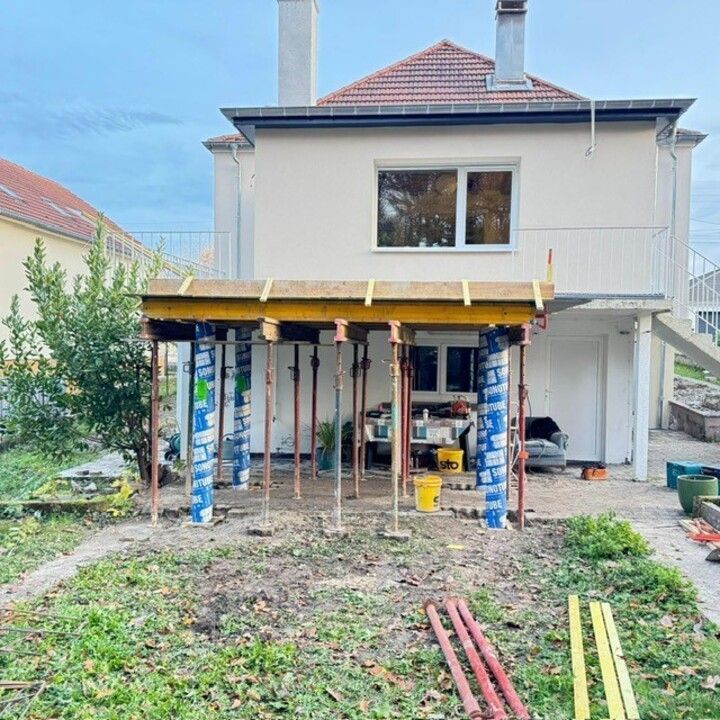 Extérieur de la maison avec un porche couvert en construction, soutenu par des poteaux peints et des outils éparpillés dans la cour