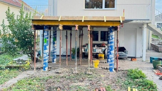 Terrasse ou patio en bois partiellement construit sous une maison, avec des poteaux de soutien