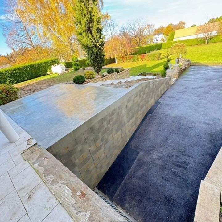 Allée extérieure longeant une terrasse surélevée en pierre, avec un jardin paysager et une pelouse verdoyante baignée de soleil.