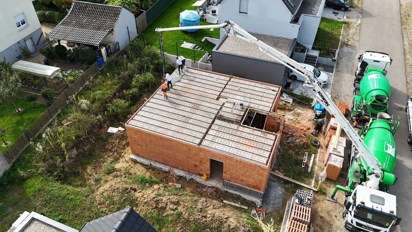 Vue aérienne d'une maison en construction avec un toit de tuiles, entourée de camions malaxeurs à béton et de matériaux de construction.