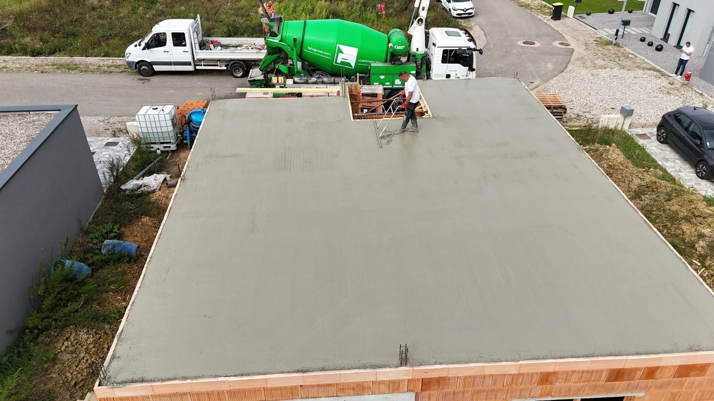 Dalle de fondation plate en béton avec un camion blanc et une bétonnière verte garés en arrière-plan près d'une allée.
