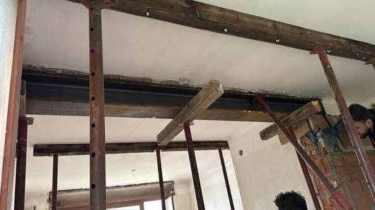 Scène de construction intérieure avec poutres de plafond apparentes et ouvriers installant une planche de bois sur un échafaudage.