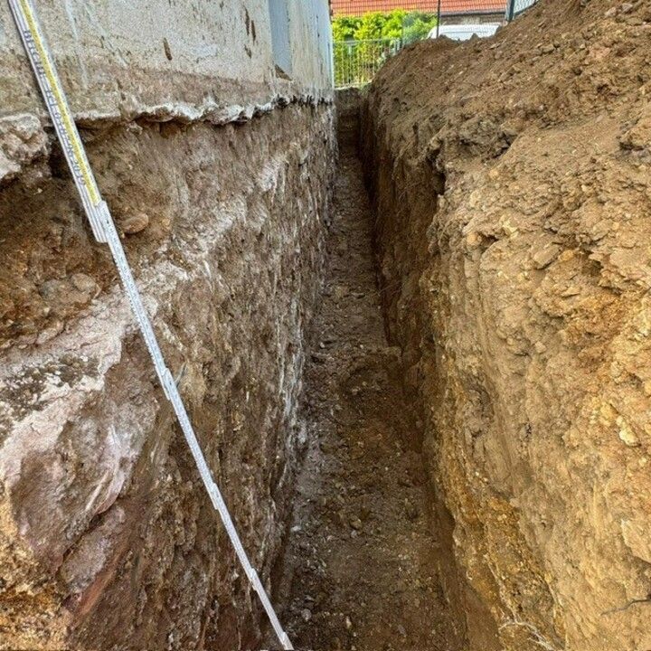 Tranchée étroite creusée le long d'un mur de bâtiment, mettant à nu le sol et les fondations.