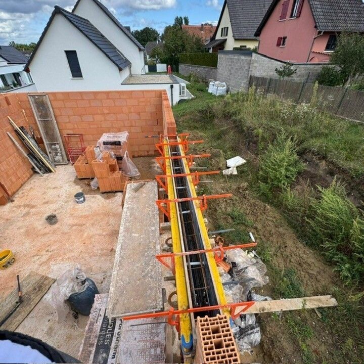 Chantier avec échafaudages orange au-dessus d'une étroite tranchée en béton à côté d'une maison et d'un mur en briques