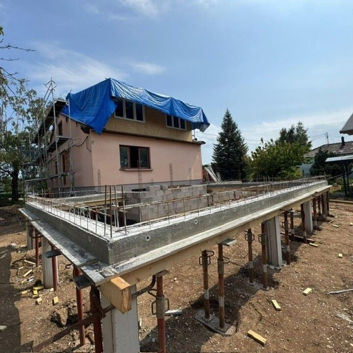 Fondations en béton d'une maison en construction à côté d'une maison rose à deux étages avec une bâche bleue sur le toit.