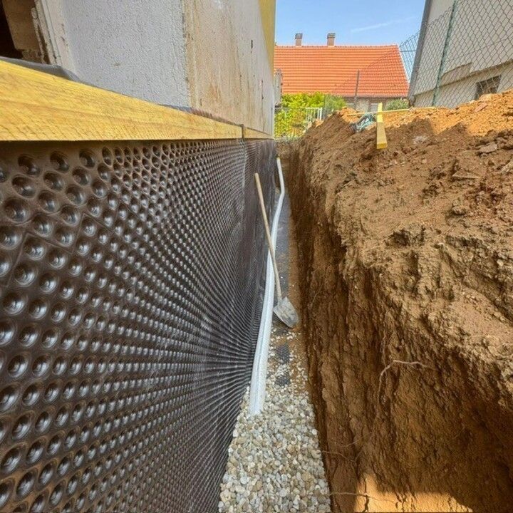 Excavation le long d'un mur de bâtiment avec membrane d'étanchéité noire et tuyau de drainage blanc dans une tranchée étroite