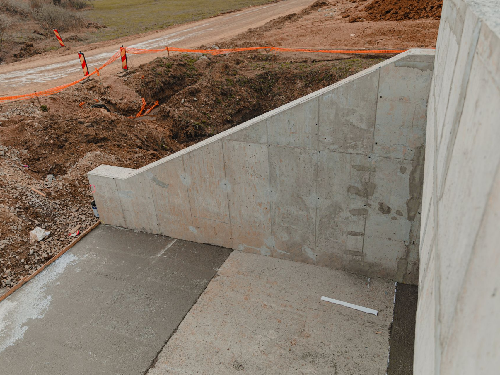 Rampe en béton et mur en pente bordant un chemin de terre balisé de bornes orange.