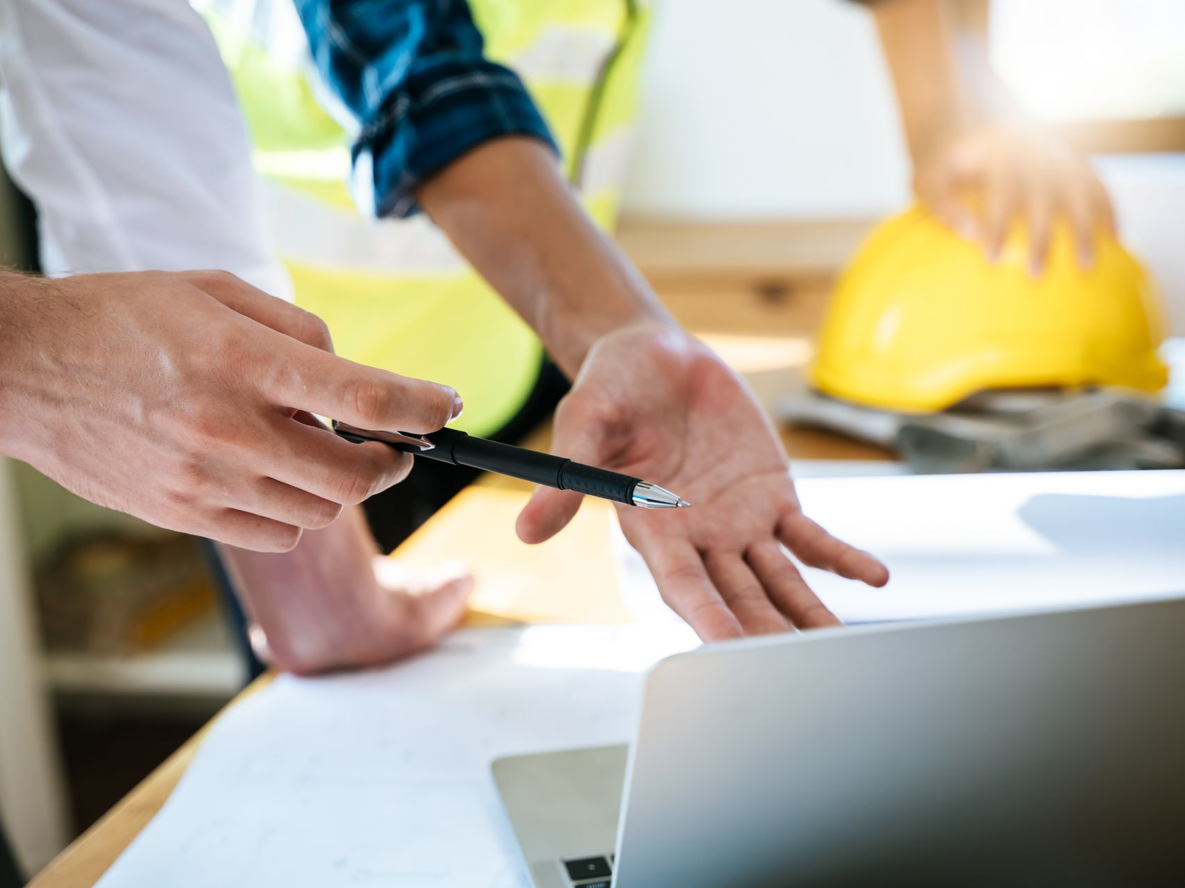 Des mains pointent un ordinateur portable lors d'une discussion d'équipe, avec des casques de chantier jaunes en arrière-plan.