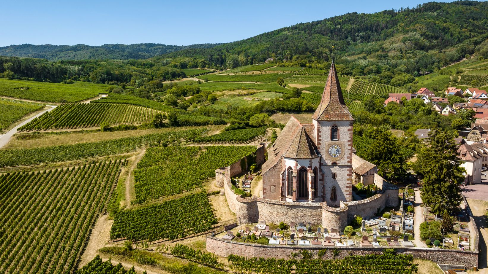 Église de pierre au milieu des vignes, dans une vallée verdoyante à flanc de colline, sous un ciel clair