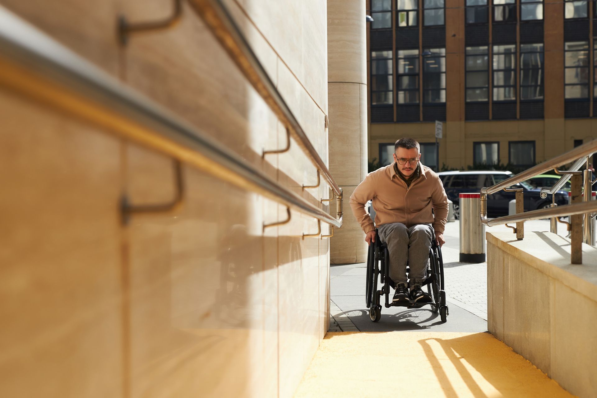 Un homme en fauteuil roulant emprunte une rampe extérieure ensoleillée entre deux bâtiments
