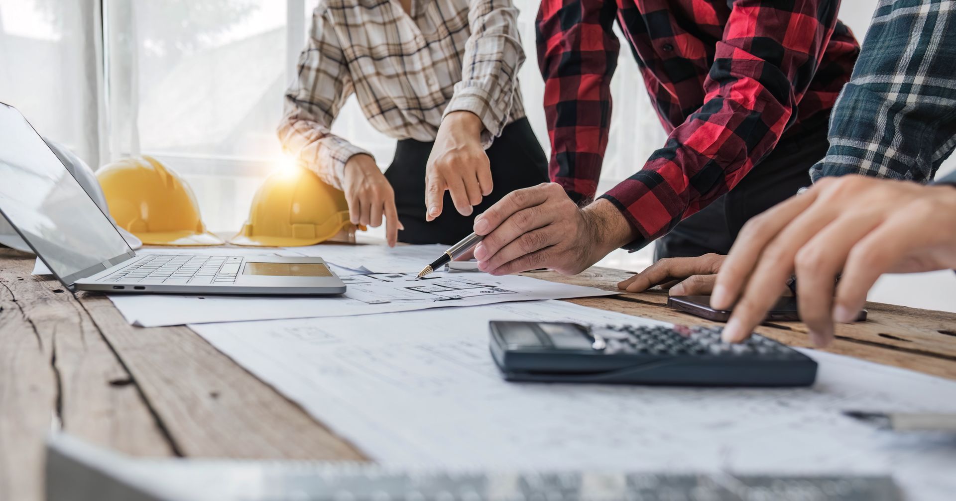 Des personnes collaborent sur des plans à un bureau équipé d'un ordinateur portable, d'une calculatrice et d'un casque de chantier.