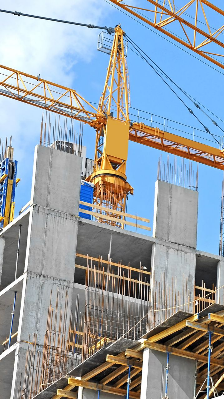 Une grue à tour jaune soulève des matériaux sur un chantier de construction avec des colonnes en béton inachevées et des échafaudages.