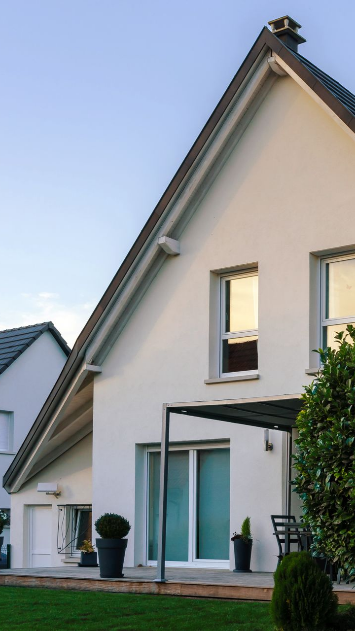 Maison blanche moderne avec un toit gris pentu, de grandes fenêtres et un petit patio couvert.