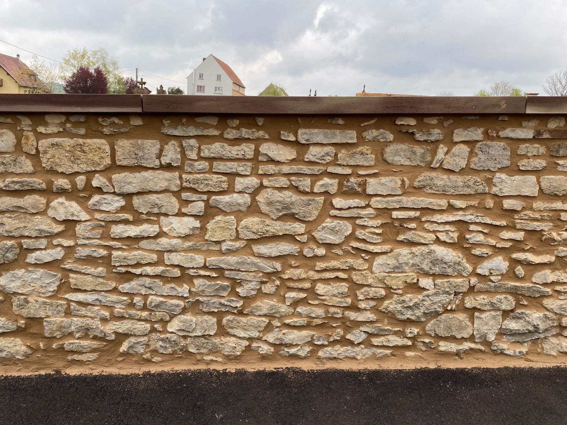Mur de pierre surmonté de bois, avec des toits et des arbres visibles au-dessus sous un ciel nuageux