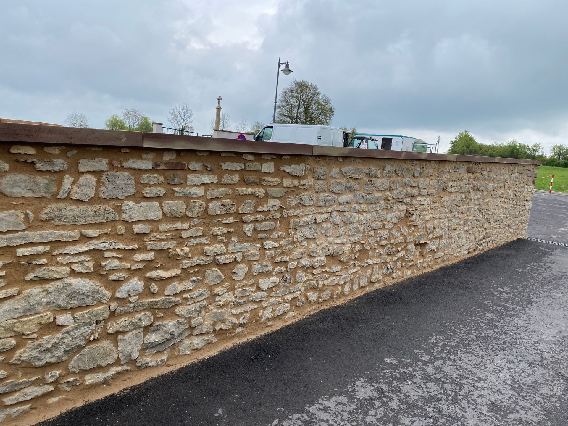 Un mur de pierre borde une route pavée sous un ciel nuageux, avec des arbres et quelques objets au loin.