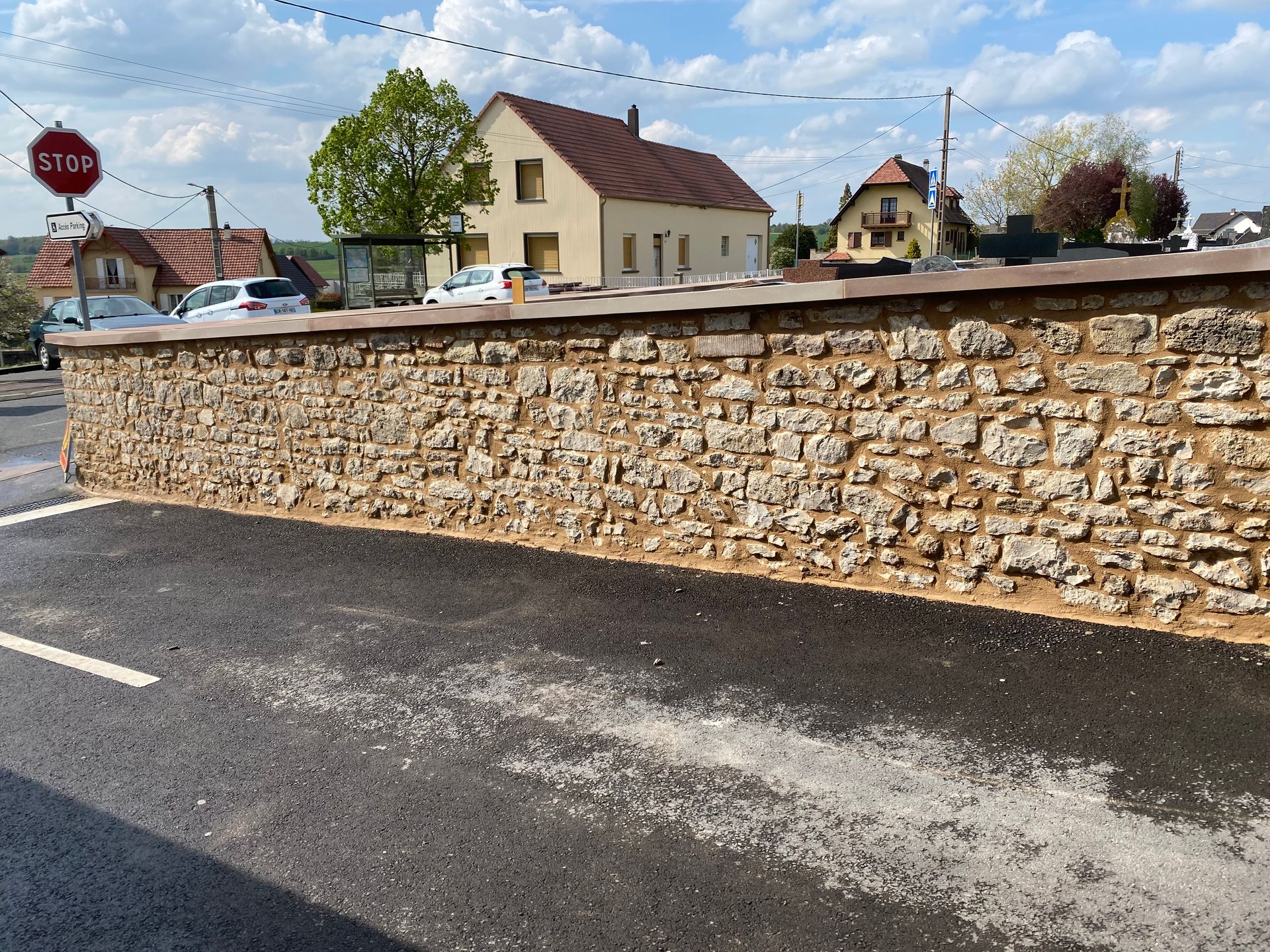 Mur en pierre bordant une route goudronnée dans un village tranquille, avec des maisons et un panneau stop en arrière-plan.