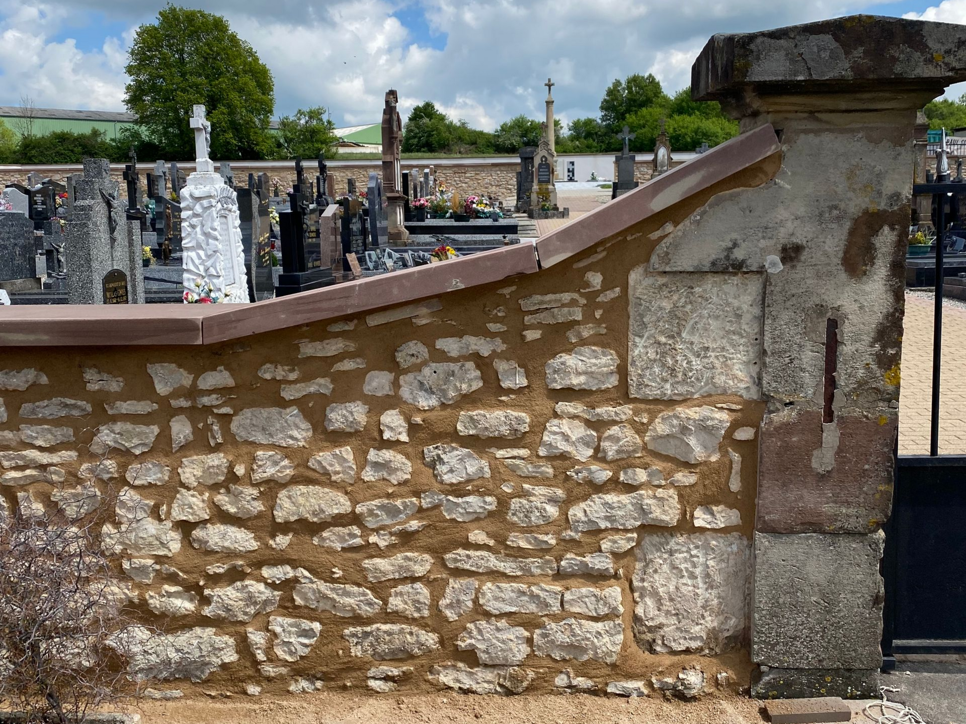Mur de pierre à sommet arrondi dans un cimetière, avec des pierres tombales et des arbres en arrière-plan