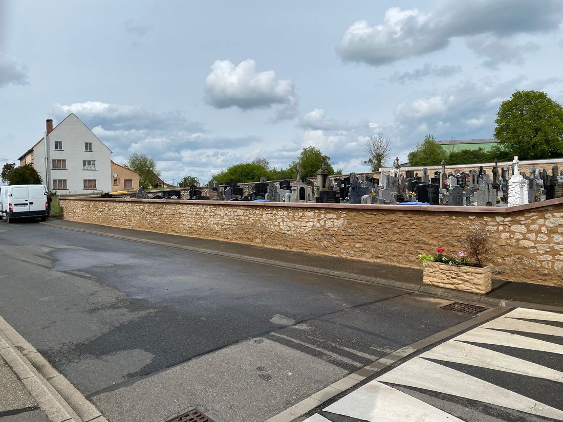 Large rue longeant un mur de pierre et un cimetière sous un ciel nuageux, avec des voitures garées et une maison blanche.