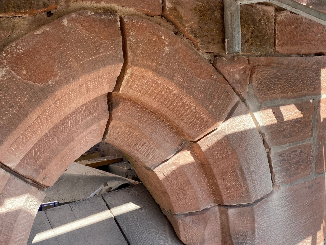 Gros plan sur une arche en briques brun rougeâtre présentant une fissure et des ombres projetées par la lumière du soleil.