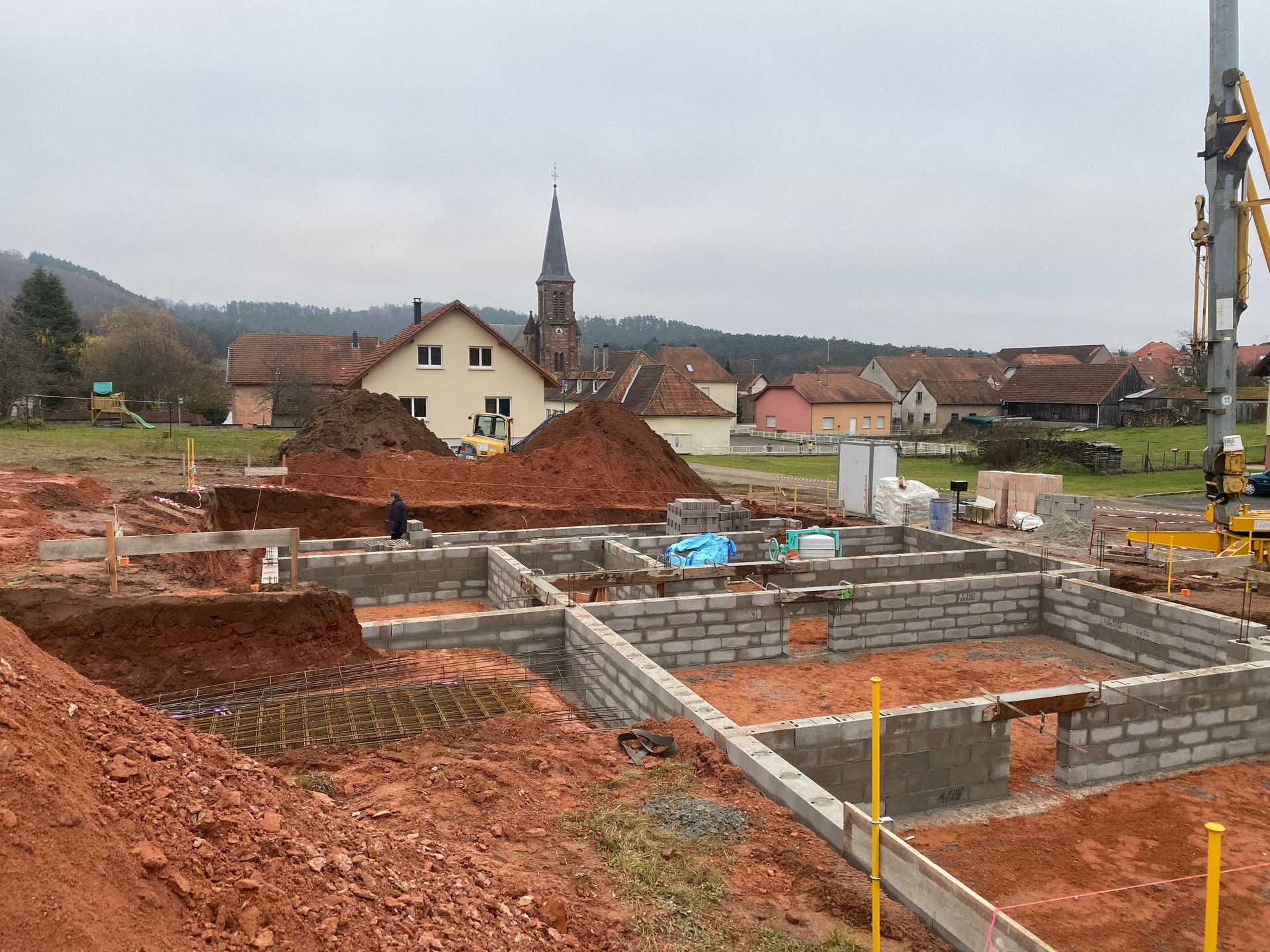 Chantier de construction avec terre rouge, fondations en béton et une grue surplombant les maisons du village et le clocher d'une église.