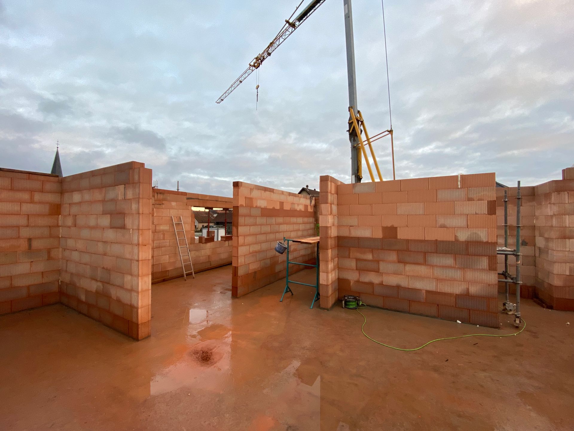 Murs d'une maison en briques partiellement construits sur un chantier boueux, avec une grue au-dessus.