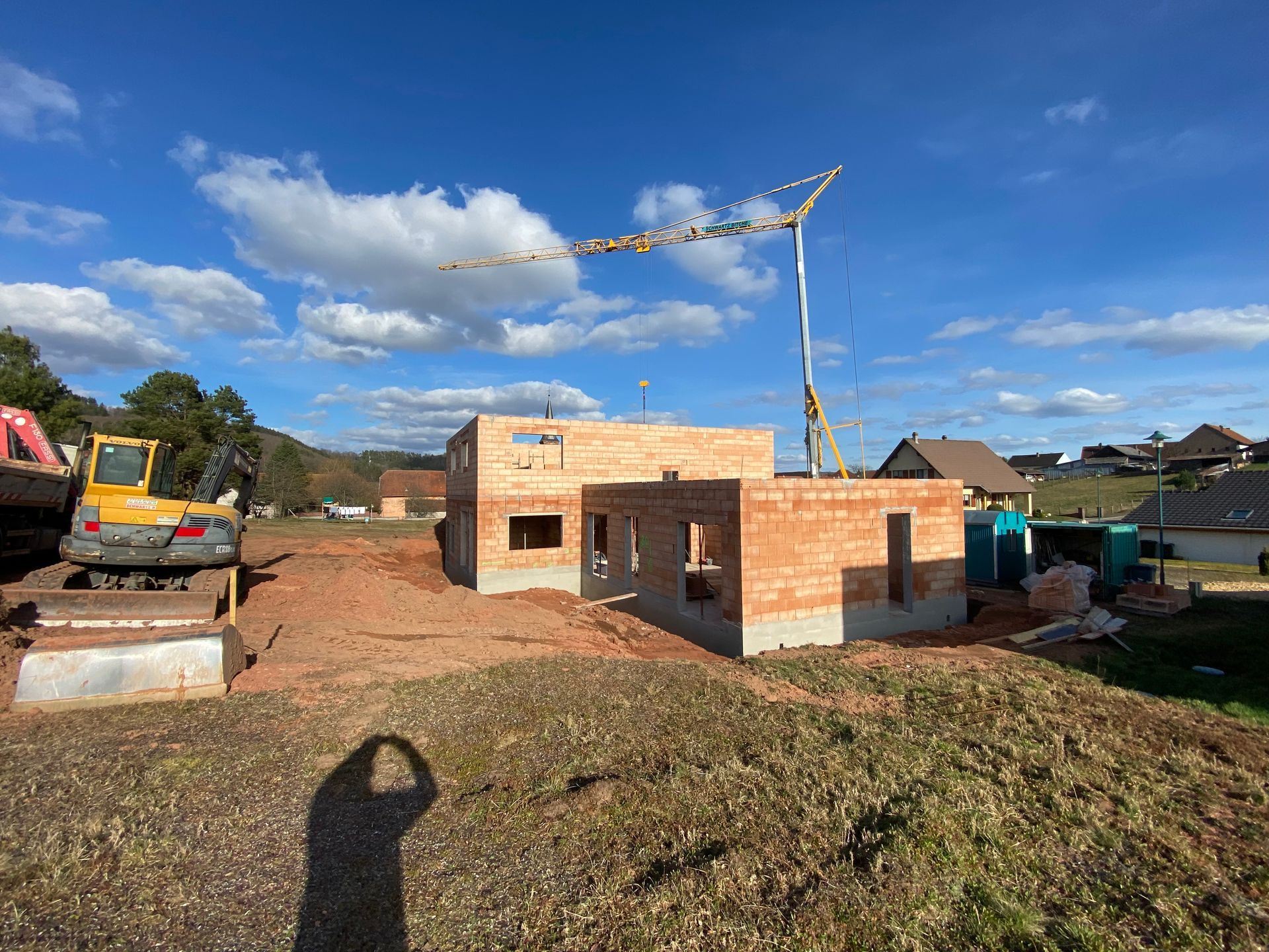 Maison en briques partiellement construite sur un terrain boueux, sous un ciel bleu, avec une grue au-dessus.