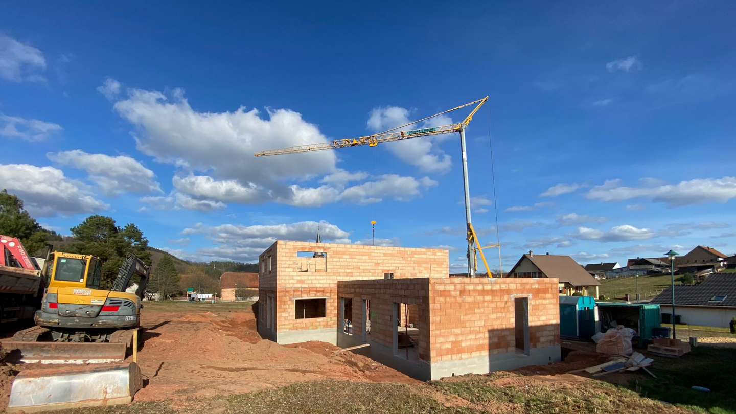 Maison en briques en construction avec une pompe à béton, une excavatrice et un ciel bleu parsemé de nuages.