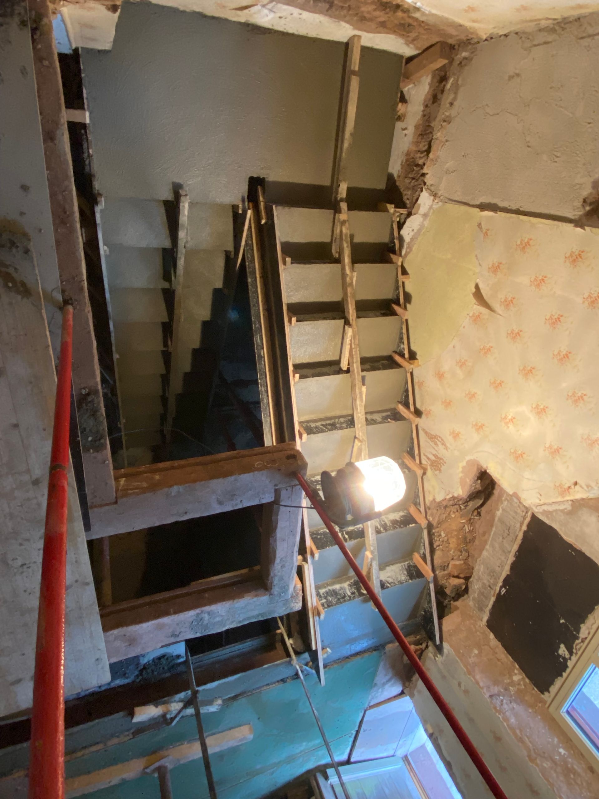 Cage d'escalier sombre avec marches en bois, murs apparents et une lampe de travail puissante éclairant un sol bleu.