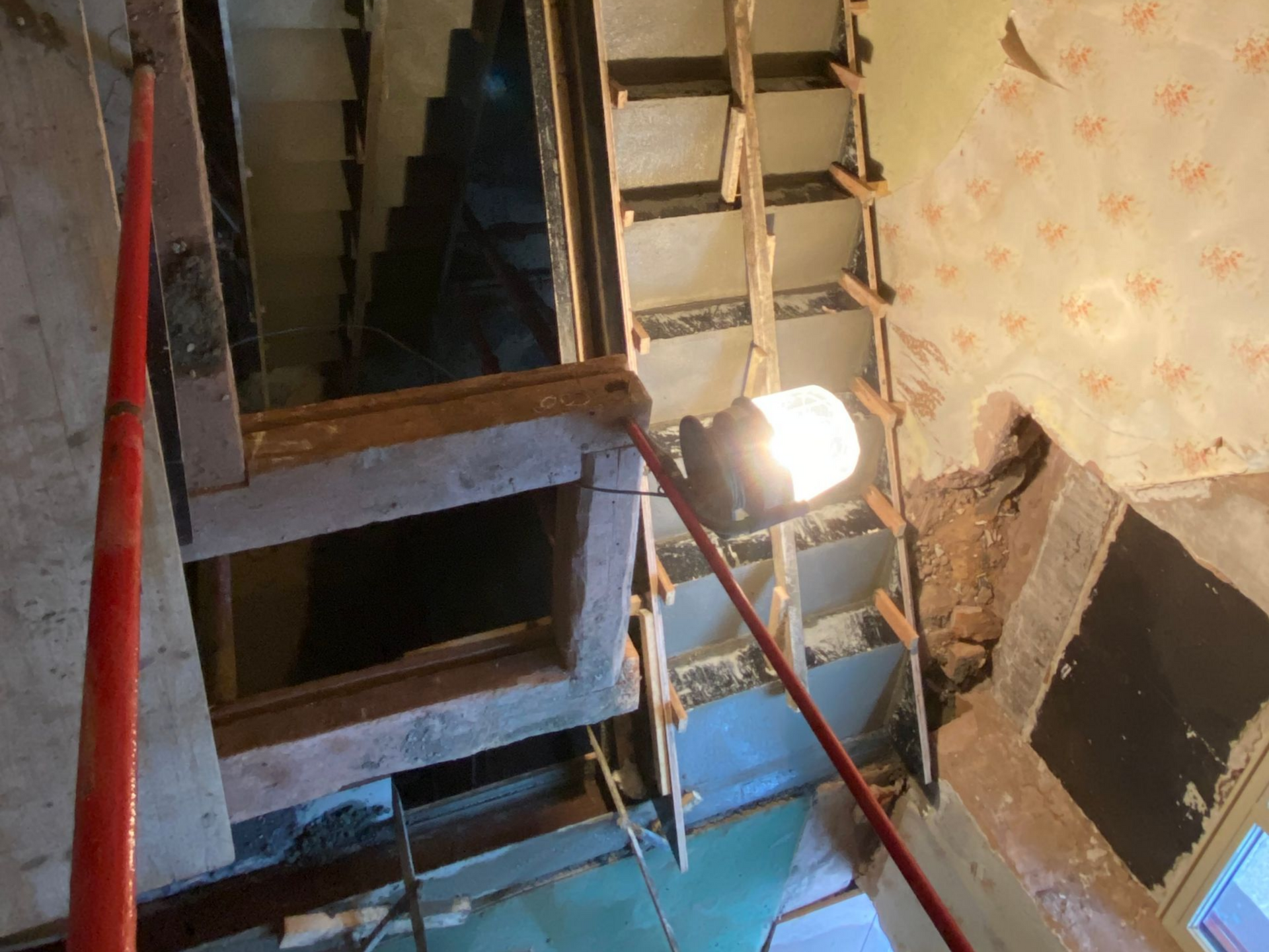 Cage d'escalier endommagée avec une échelle, une charpente apparente et une lampe de travail puissante dans un espace intérieur exigu.