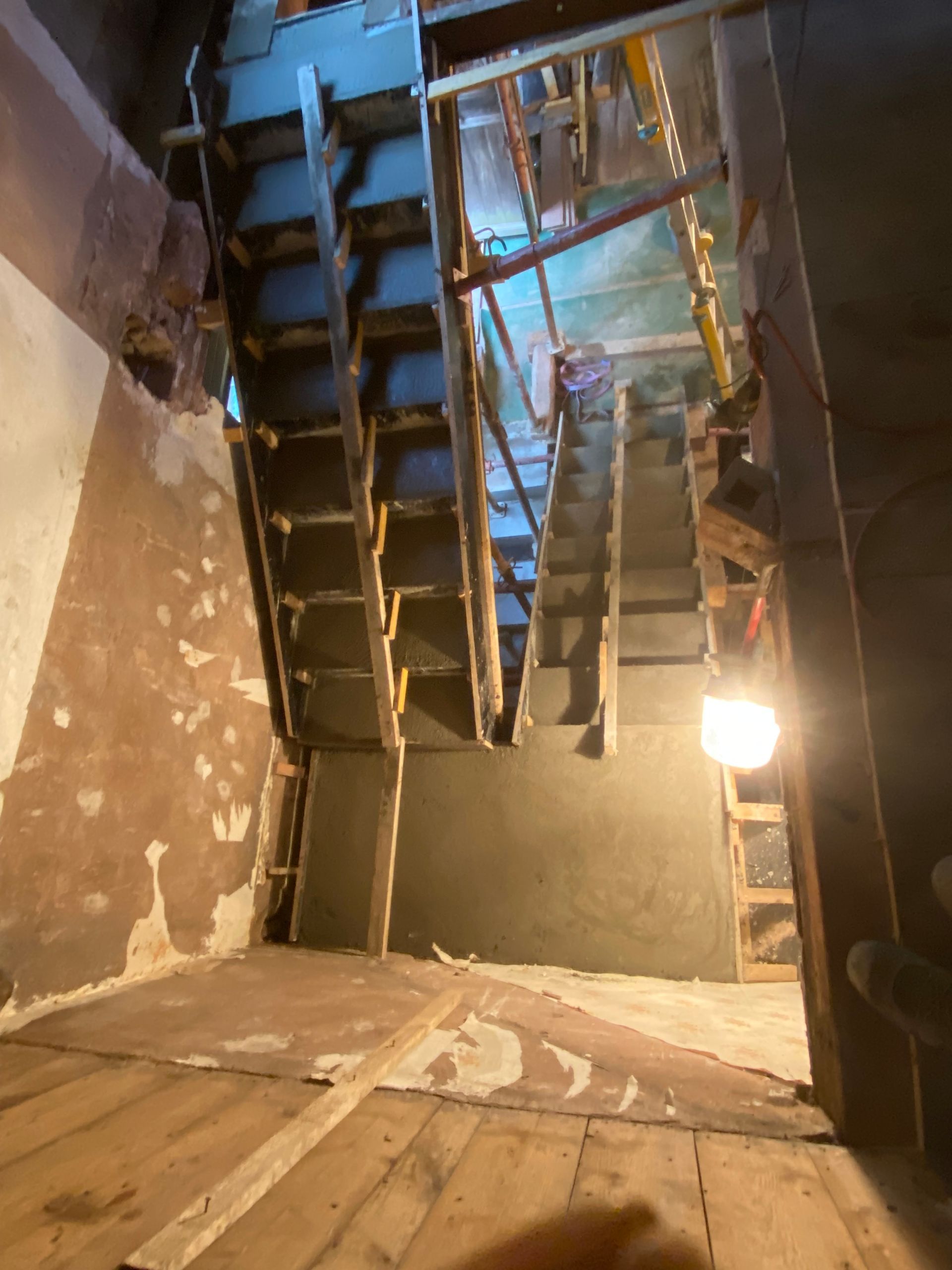 Escalier intérieur endommagé avec poutres apparentes, murs qui s'écaillent et une ampoule allumée dans une cage d'escalier étroite.