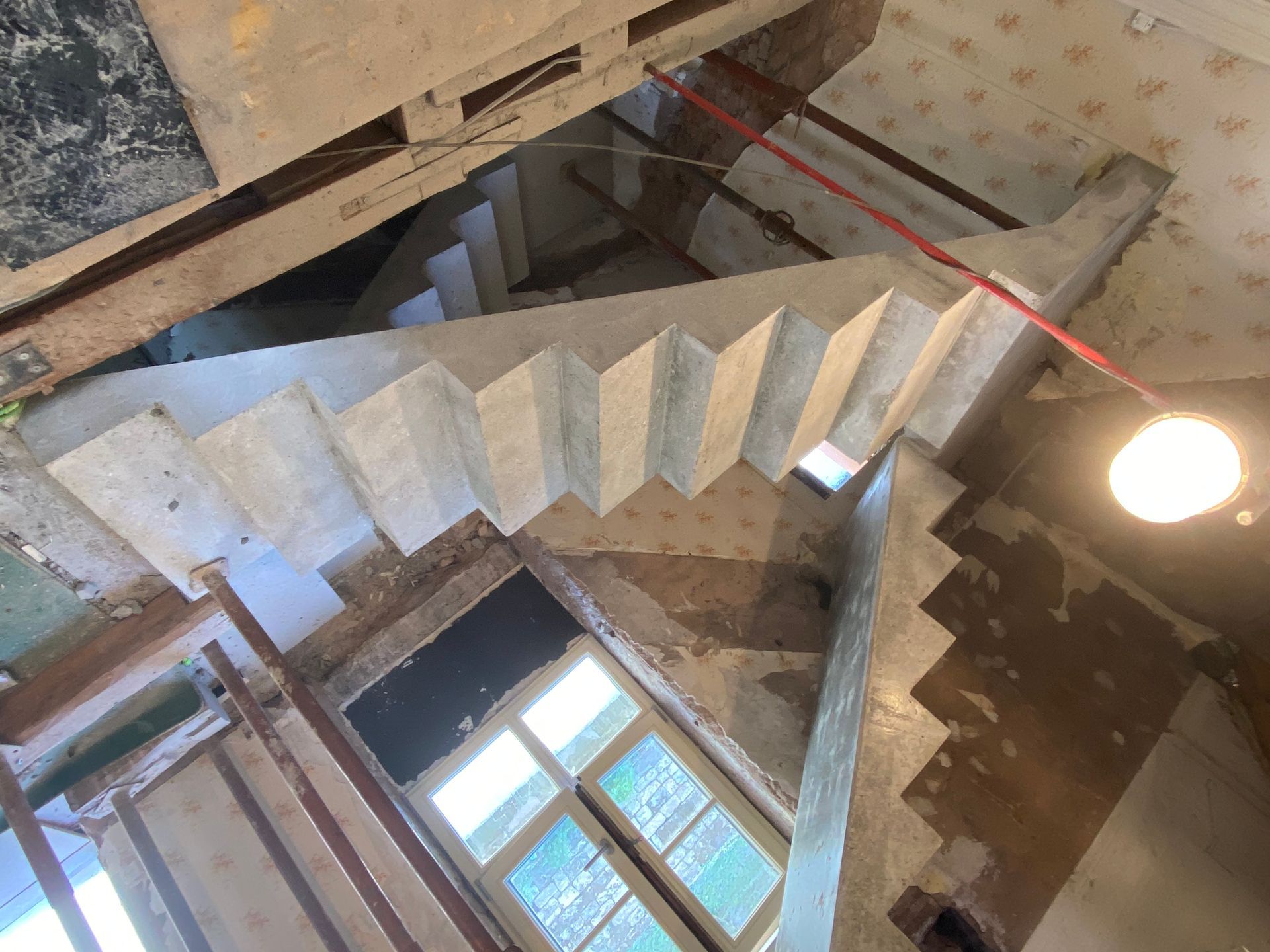 Escalier en colimaçon en béton vu de dessus, avec une fenêtre et un plafonnier apparent en dessous.