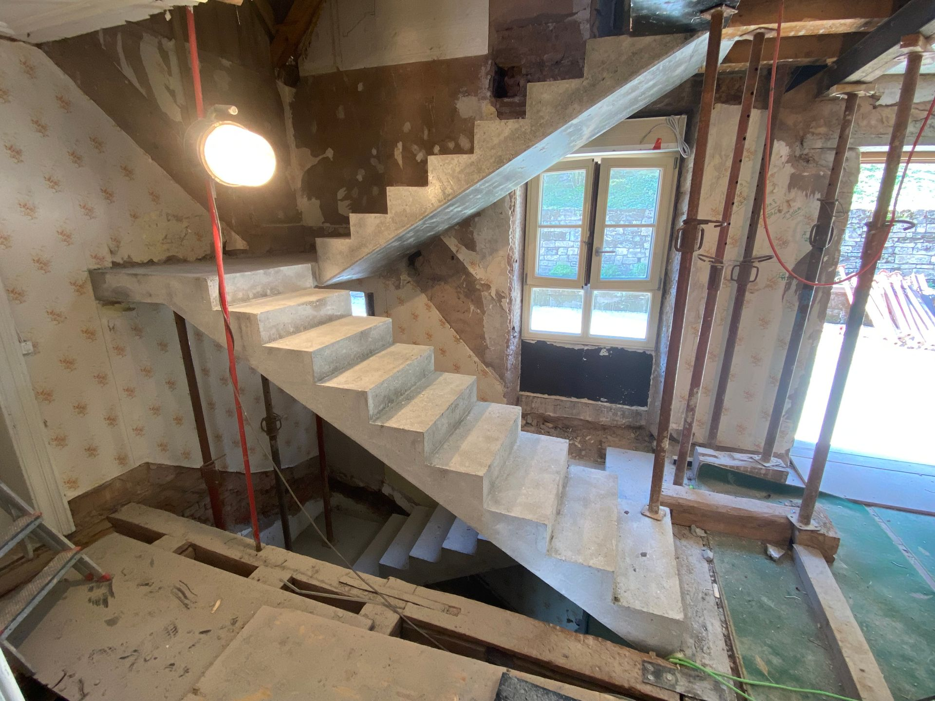 Intérieur en travaux avec escalier en béton inachevé et échafaudages près d'une fenêtre et d'une porte ouverte