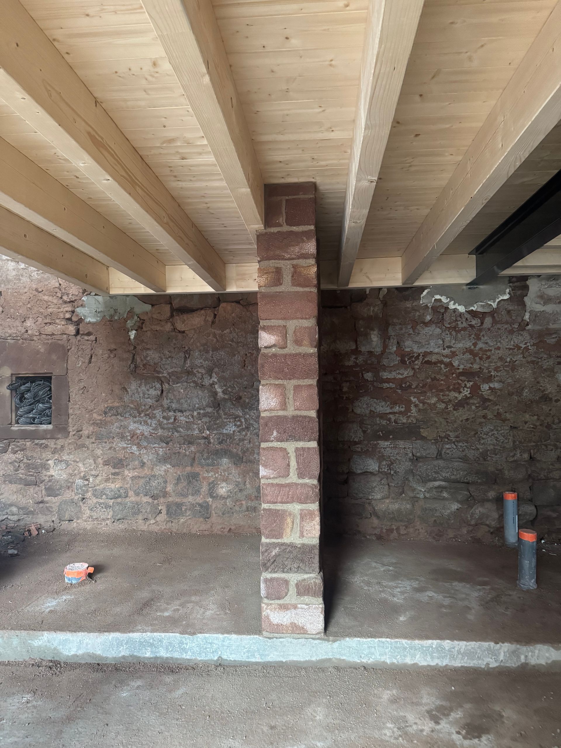 Sous-sol en construction avec poutres apparentes au plafond, pilier de soutien en briques et murs en briques non finis