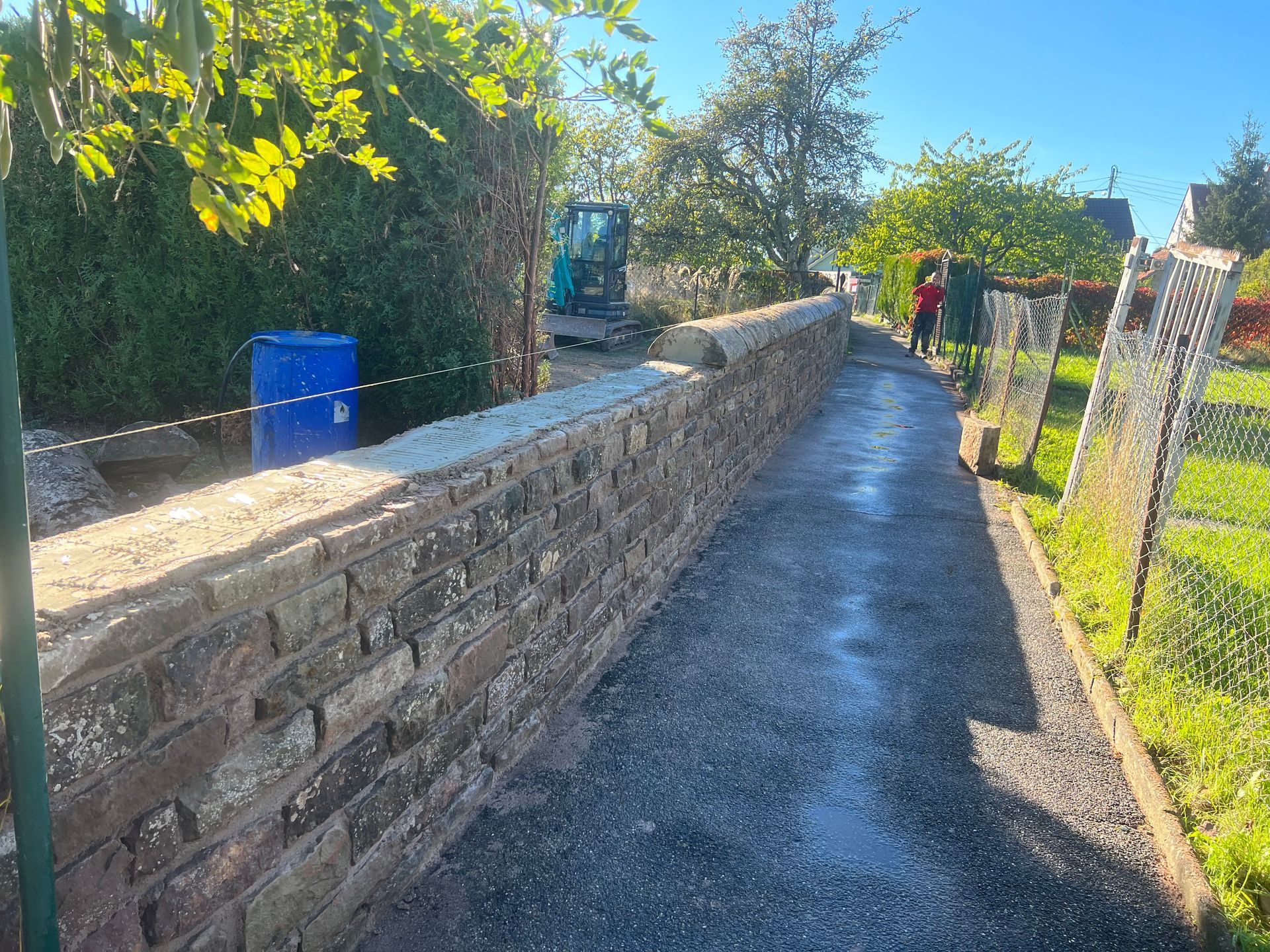 Étroit chemin pavé longeant un muret et une clôture en pierre, avec une poubelle bleue et de la verdure baignée de soleil.