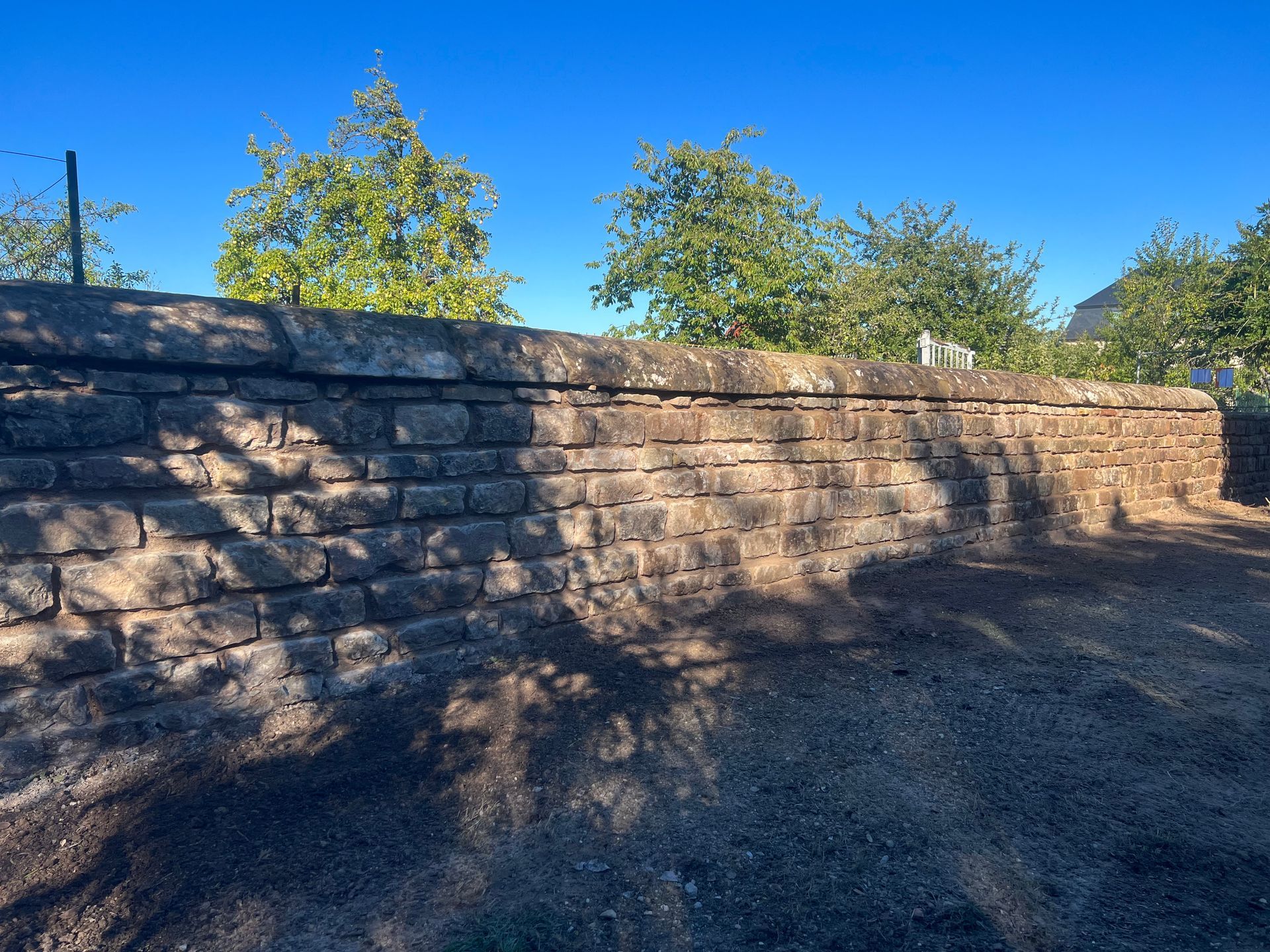 Mur de soutènement en pierre baigné de soleil, avec des arbres et un ciel bleu au-dessus, projetant de longues ombres en dessous.