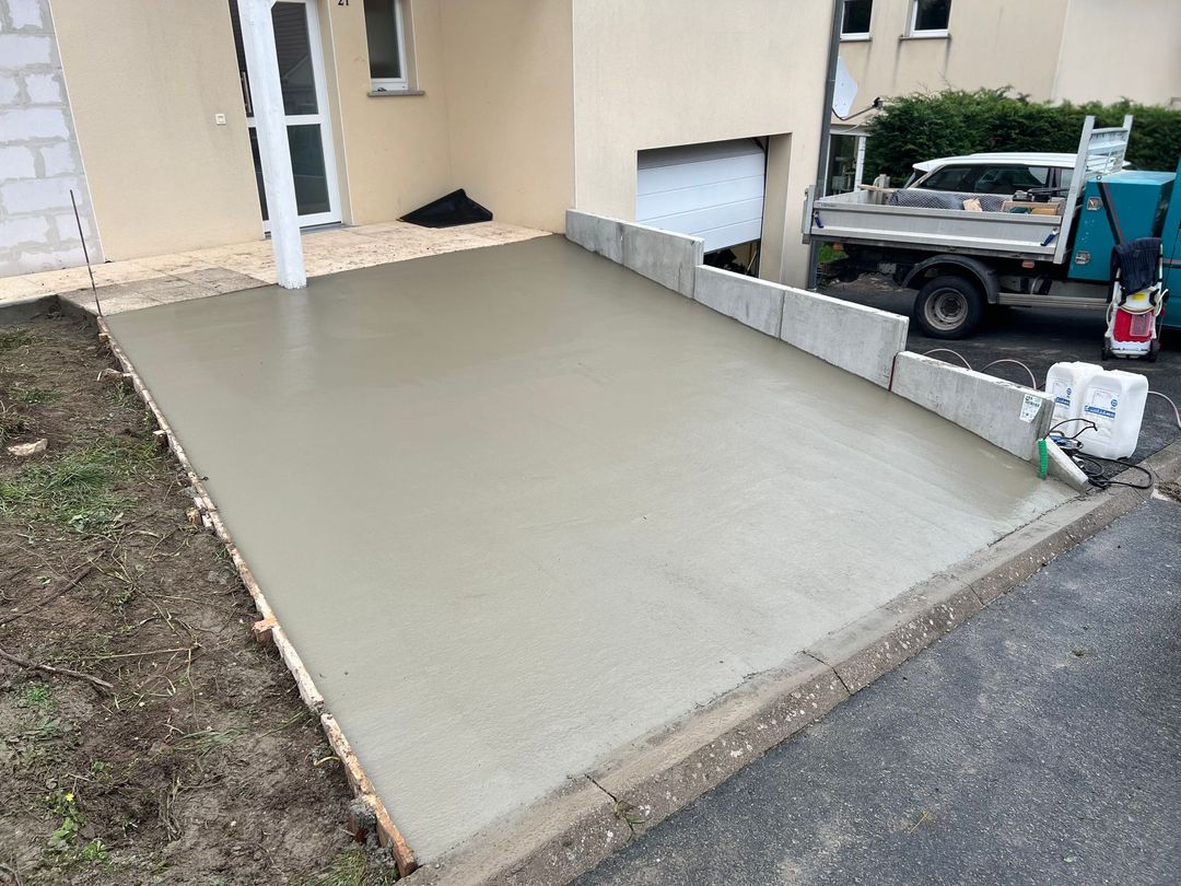 Allée en béton fraîchement coulée à côté d'une maison beige, avec une section en pente douce et un camion stationné à proximité.