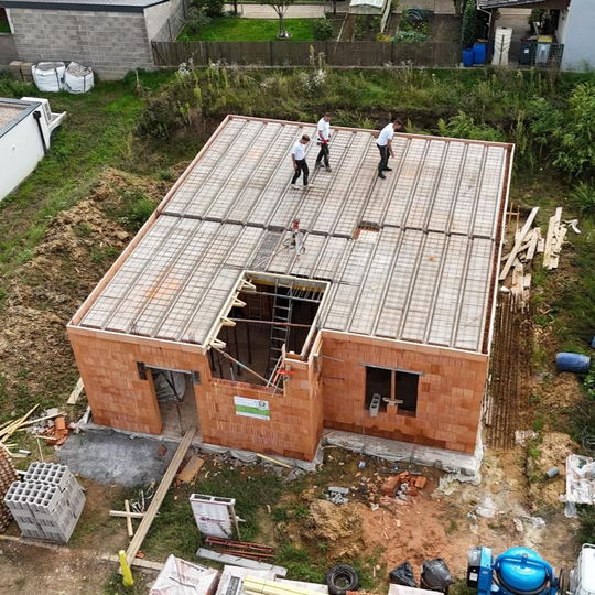 Des ouvriers travaillent sur le toit d'une maison en briques en construction, la charpente étant apparente et des matériaux étant visibles autour du chantier.