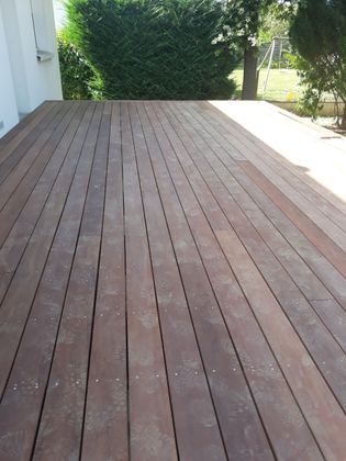 Une terrasse en bois aux planches parallèles s'étendant d'un bâtiment vers une haie et des arbres par une journée ensoleillée.