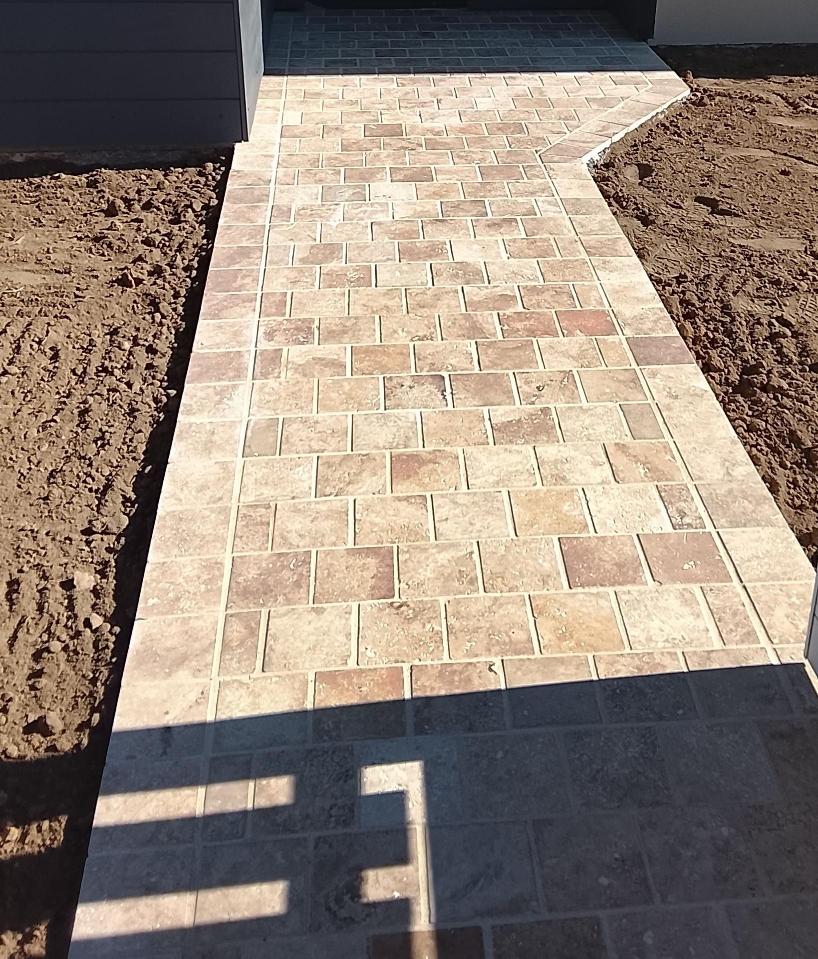 Une allée pavée de pierre beige mène à l'entrée d'une maison, bordée de zones de terre nue.